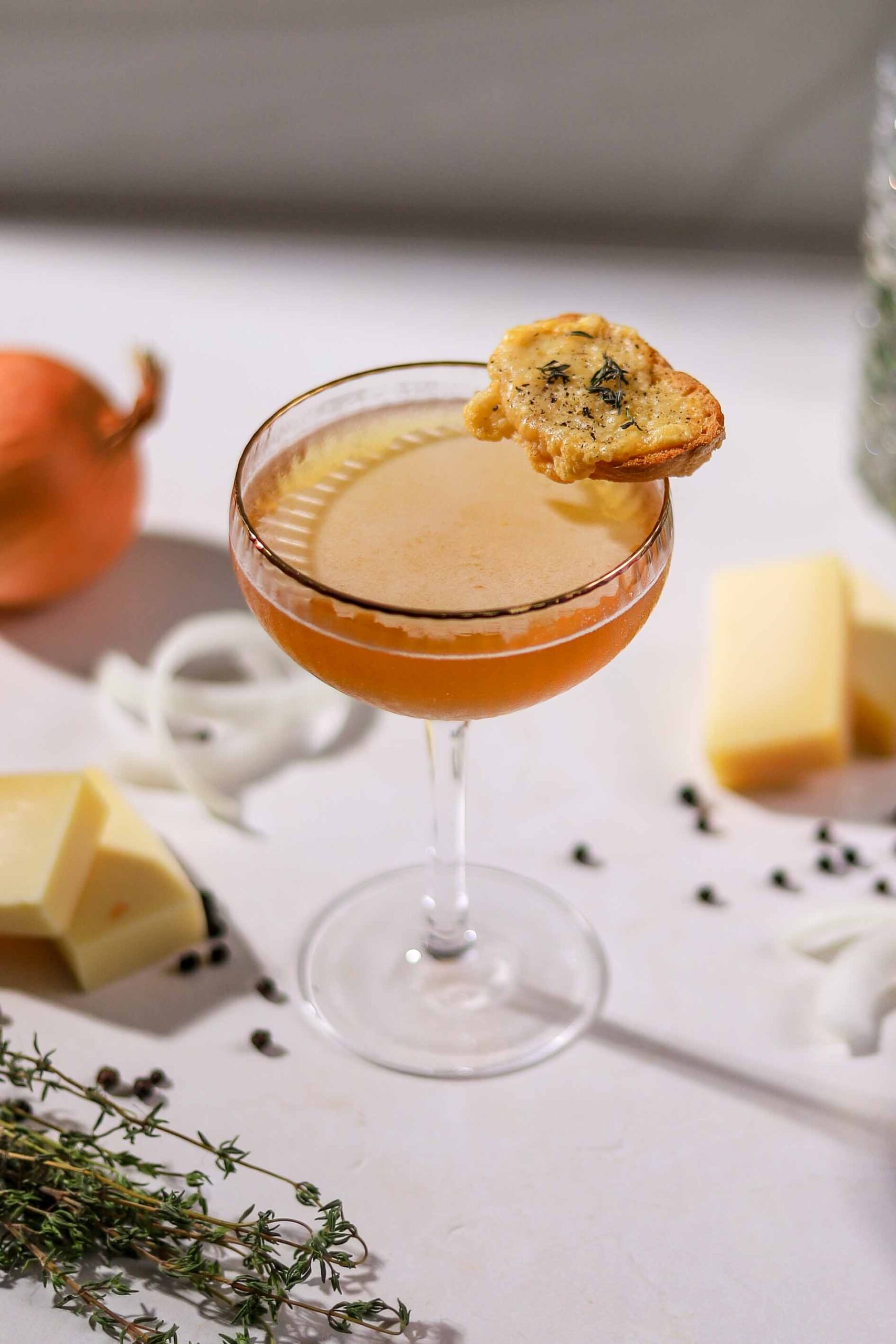 French onion soup martini in a coupe glass garnished with a Gruyère toast, with onions, thyme and cheese on the side