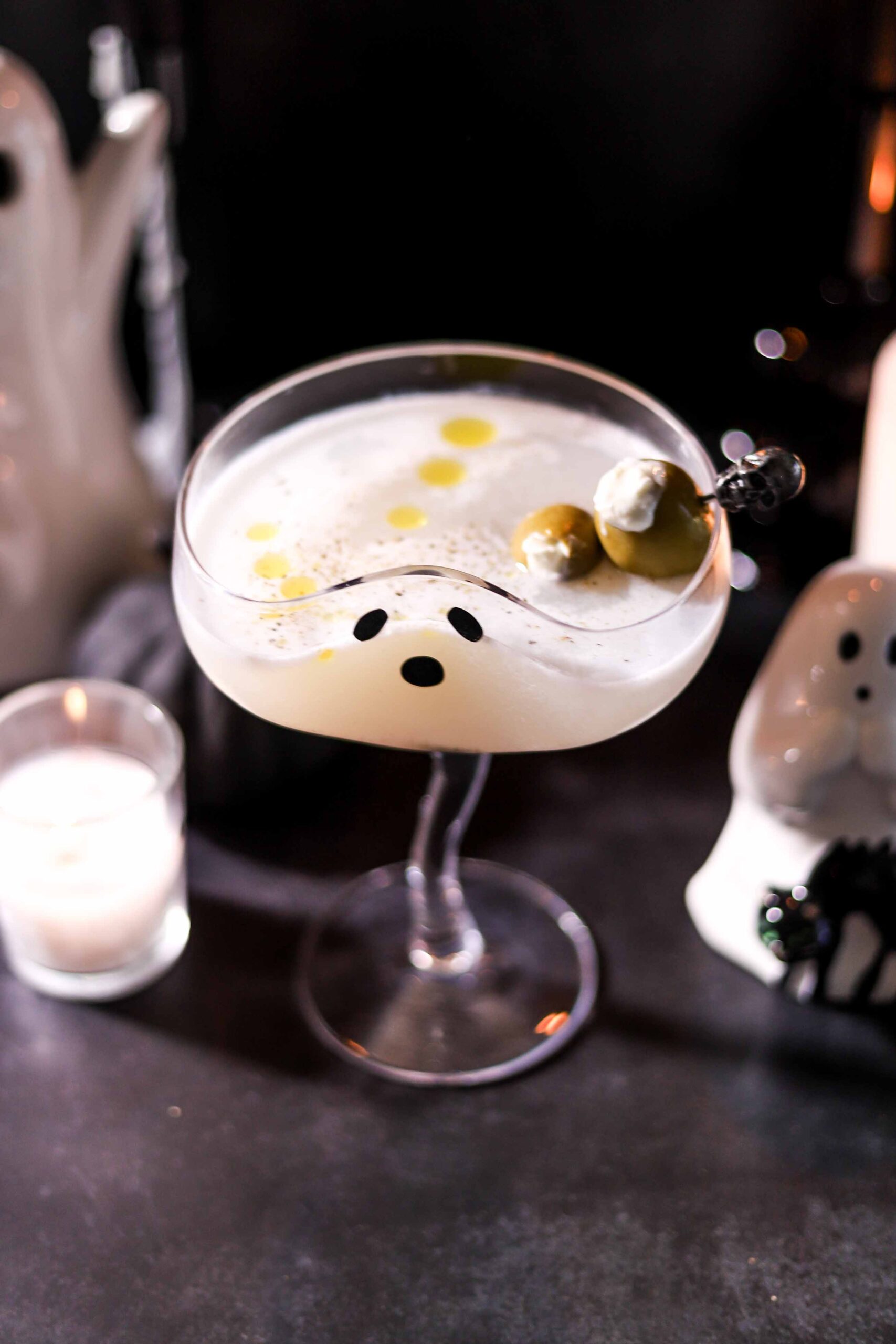 Burrata martini in a coupe glass with a ghost face, garnished with burrata-stuffed Castelvetrano olives on a pick