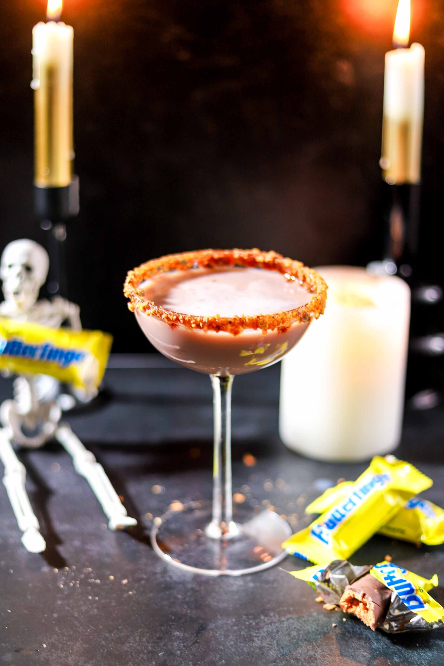 Butterfinger martini with a crushed candy rim in a coupe glass, set between lit candles with a skeleton and Butterfinger bars
