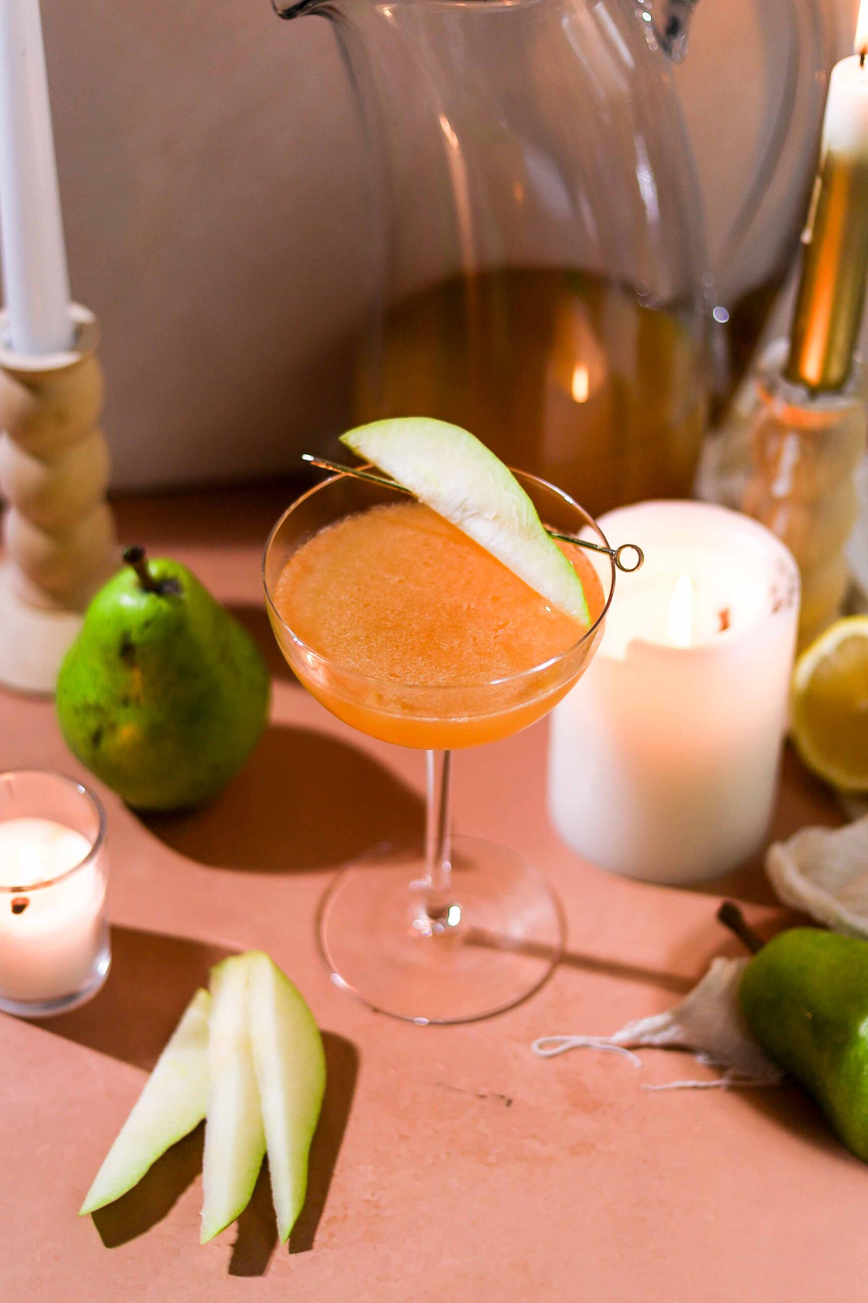 Spiced pear sidecar in a coupe glass garnished with a fresh pear slice, with a whole pear and candles in the background